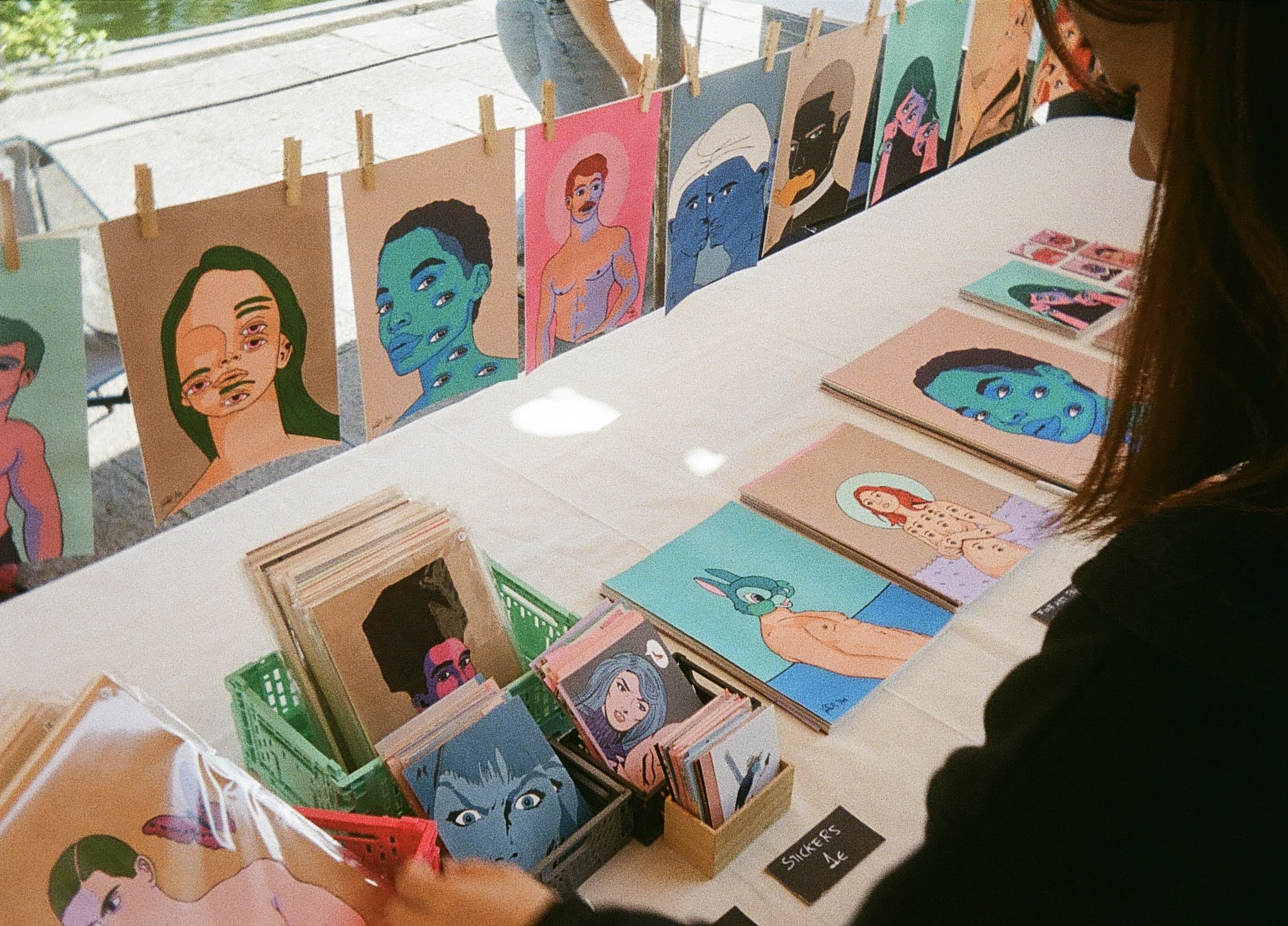 A woman looks through an aesthetically placed clothes line of colorful prints showcasing abstract faces and heads with several eyeballs and eyebrows.