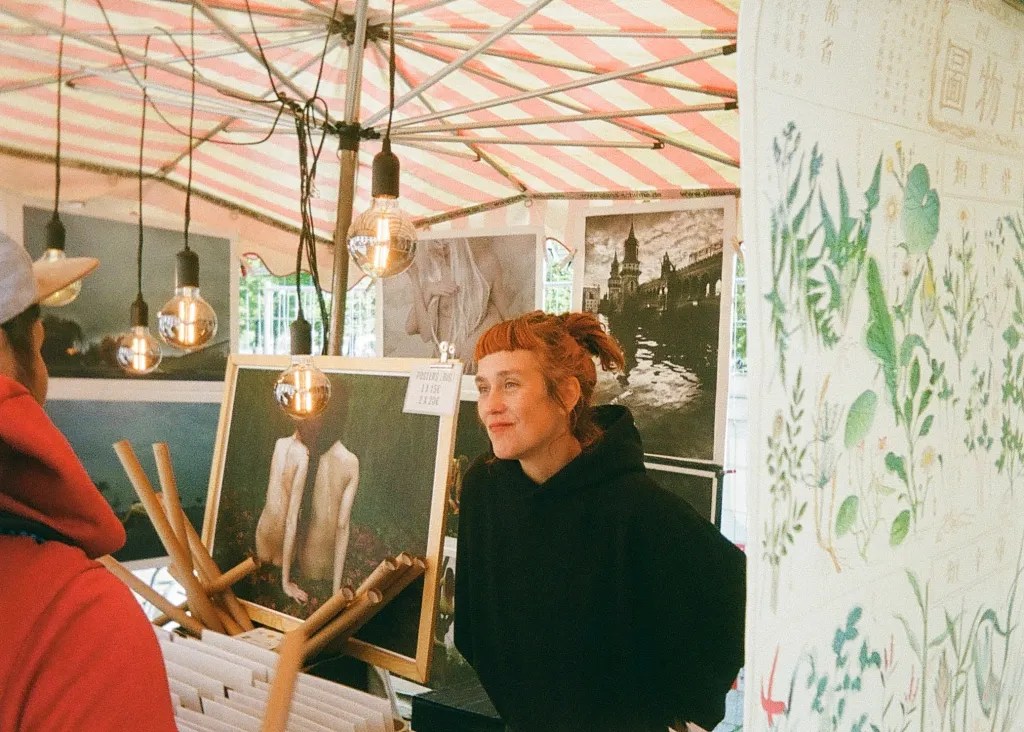 A woman at an art fair smiles at a patron, with print photographs on display in the background.
