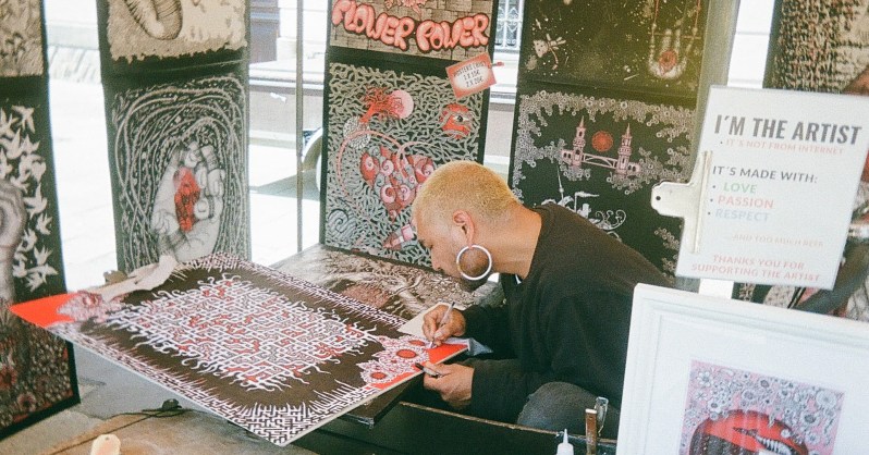A person hunches over and works on a red and black poster design, surrounded by other works of art in the background.
