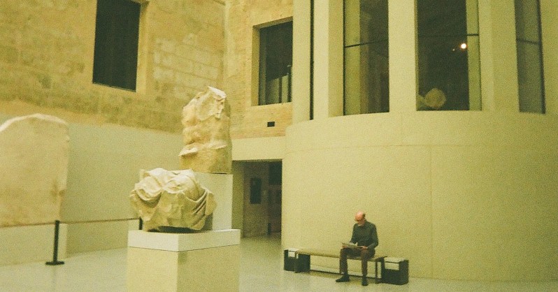The reconstructed New Museum from the outside, a man sits on a bench and two ambiguous sculptures sit outside the building.